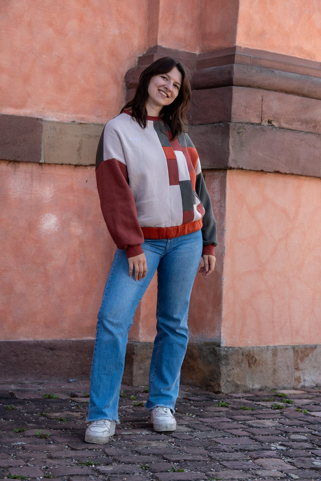 Der Boxy Sweater in Rost, Creme und Grau vor einer korallenroten Wand