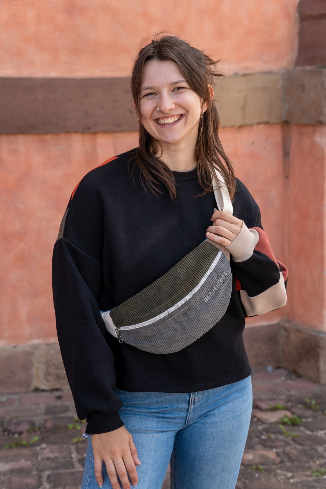 Marina lacht vor einer roten Sandsteinmauer, in einem Patchwork-Pullover und mit einer olivgrünen Koala-Bauchtasche aus Cord