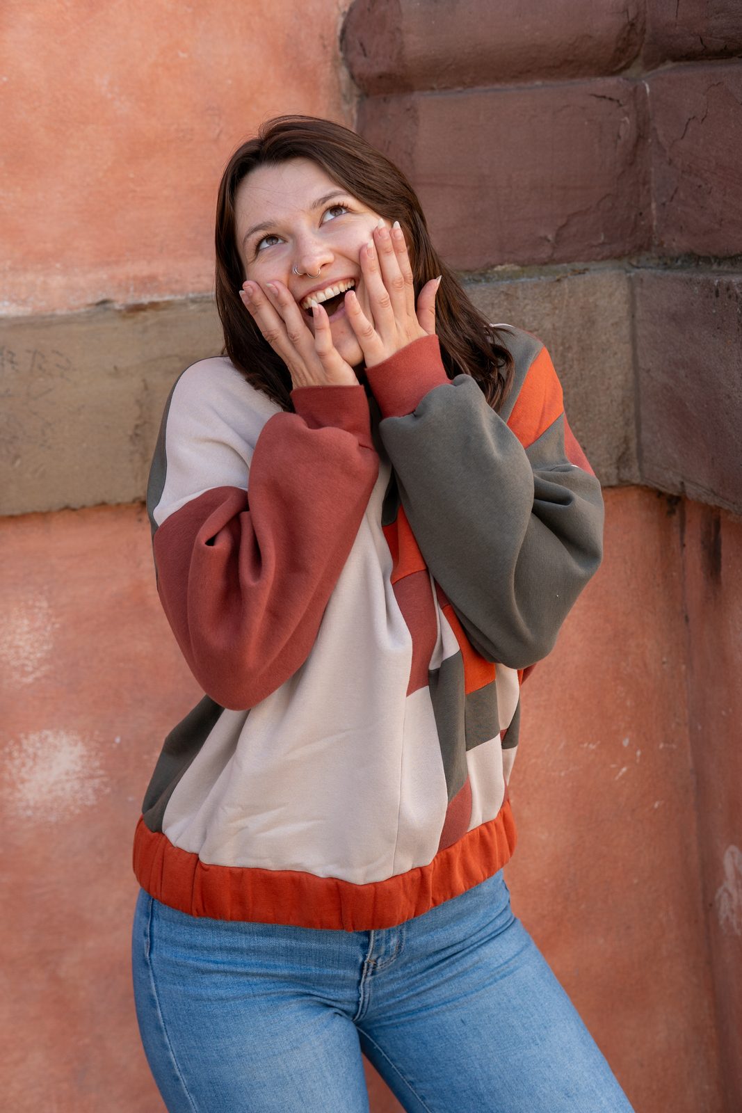 Der Boxy Sweater in Rost, Creme und Oliv, die Hände am Gesicht