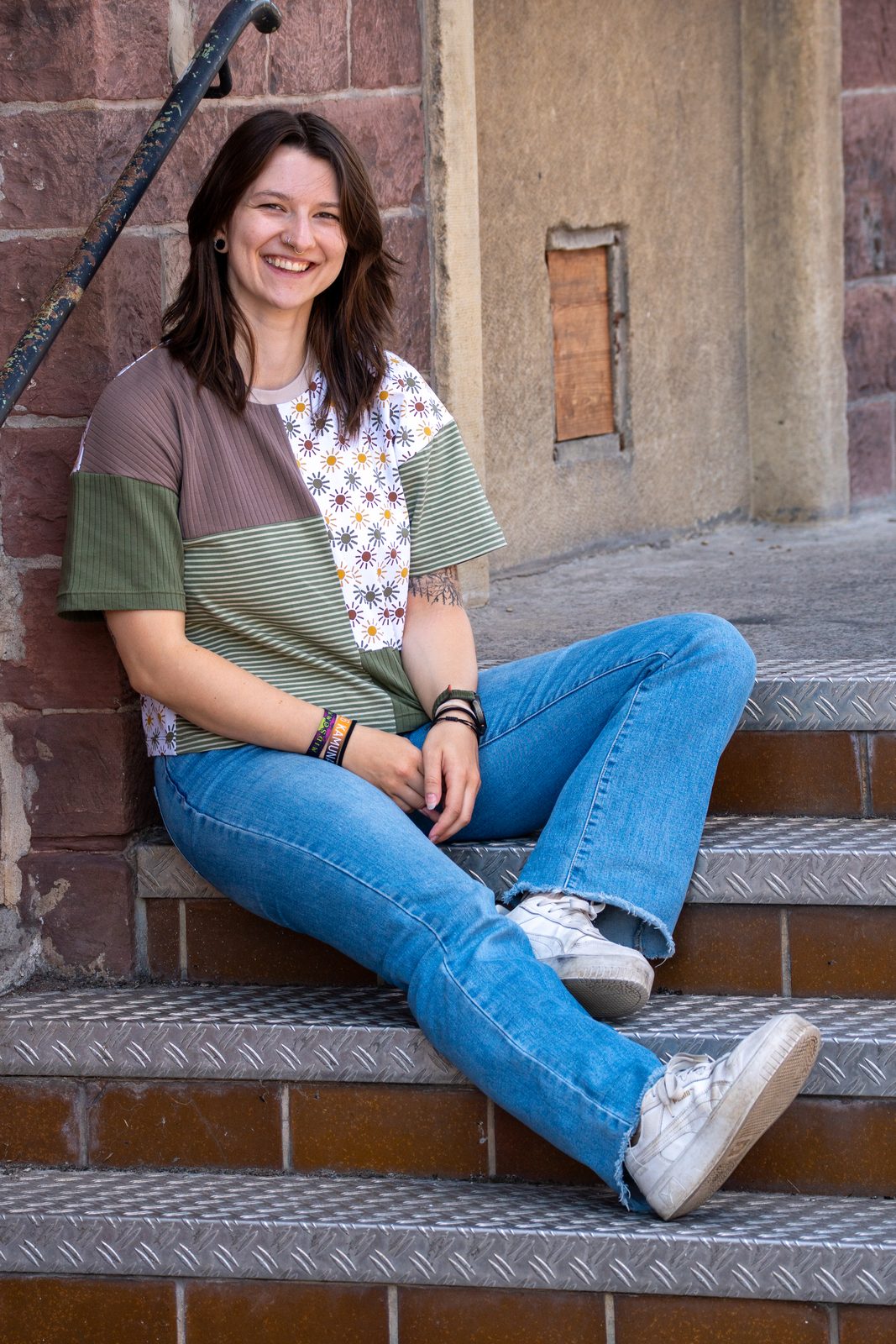 Rita in Oliv und Taupe, auf einer Metalltreppe sitzend