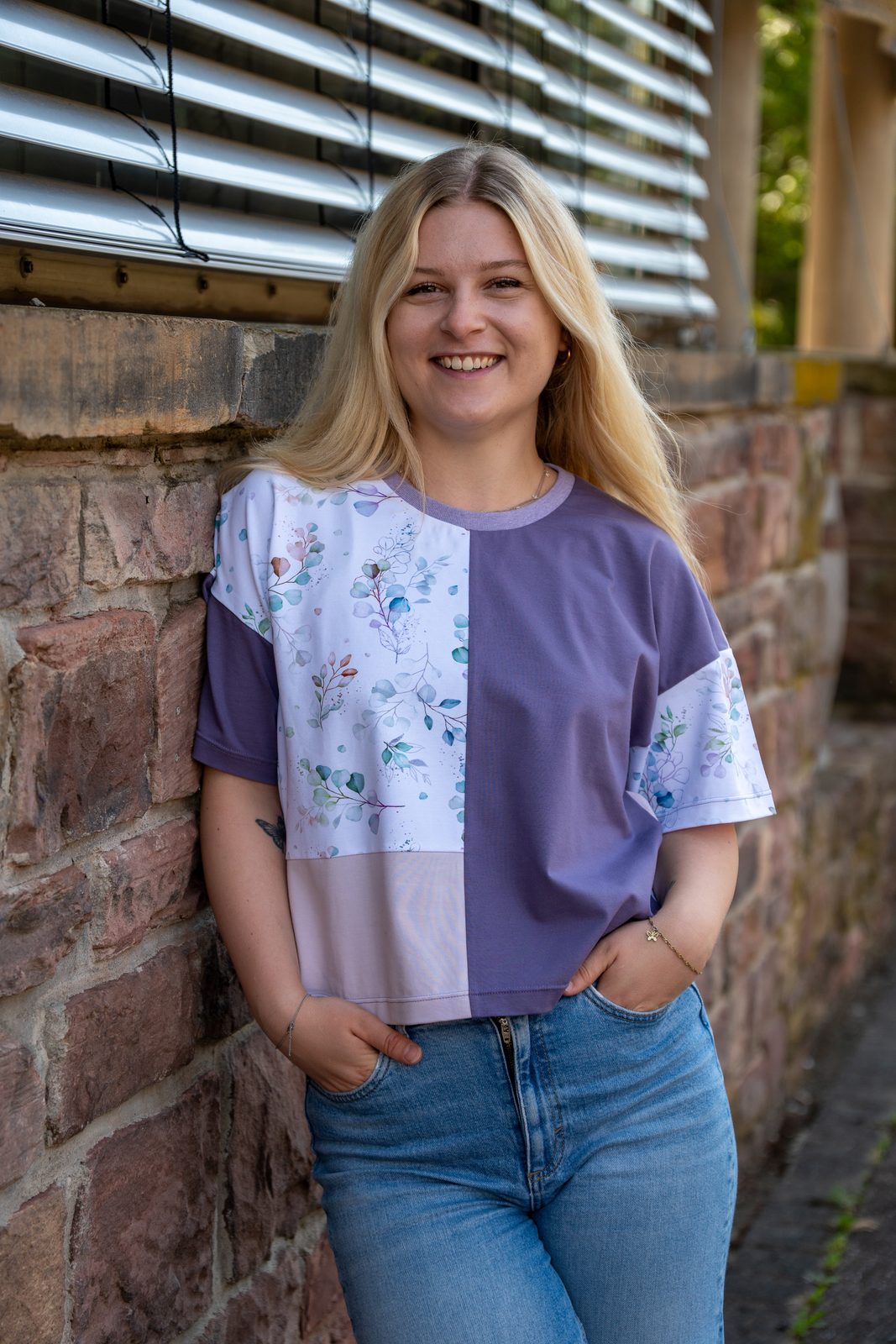 Rita in Violett, Flieder und Blumenmuster vor einer Steinwand